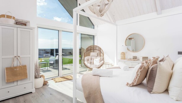 A modern bedroom with a large bed, a wicker chair, a glass door leading to a balcony, and a view of the outdoors. - No.36 South Beach - Donnini Apartments  (Troon)