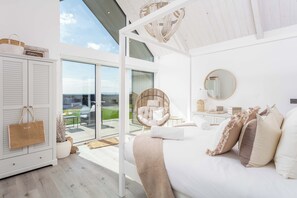 Et moderne soveværelse med en stor seng, en fletstol, en glasdør til en balkon og udsigt til det fri.