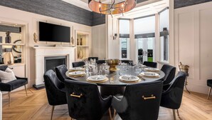 A dining room with a round table set for six, black upholstered chairs, a fireplace, a flat-screen TV mounted on the wall, and a large window offering a view of the outdoors. - No.36 South Beach - Donnini Apartments  (Troon)