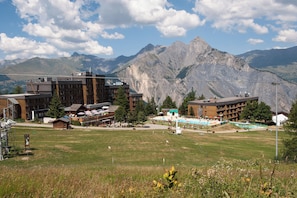 Un hotel con varias edificaciones, incluyendo una construcción de madera grande y un edificio más pequeño con techo verde. Hay una zona de piscina con cubierta verde y sillas de descanso. El hotel está rodeado de césped y árboles.