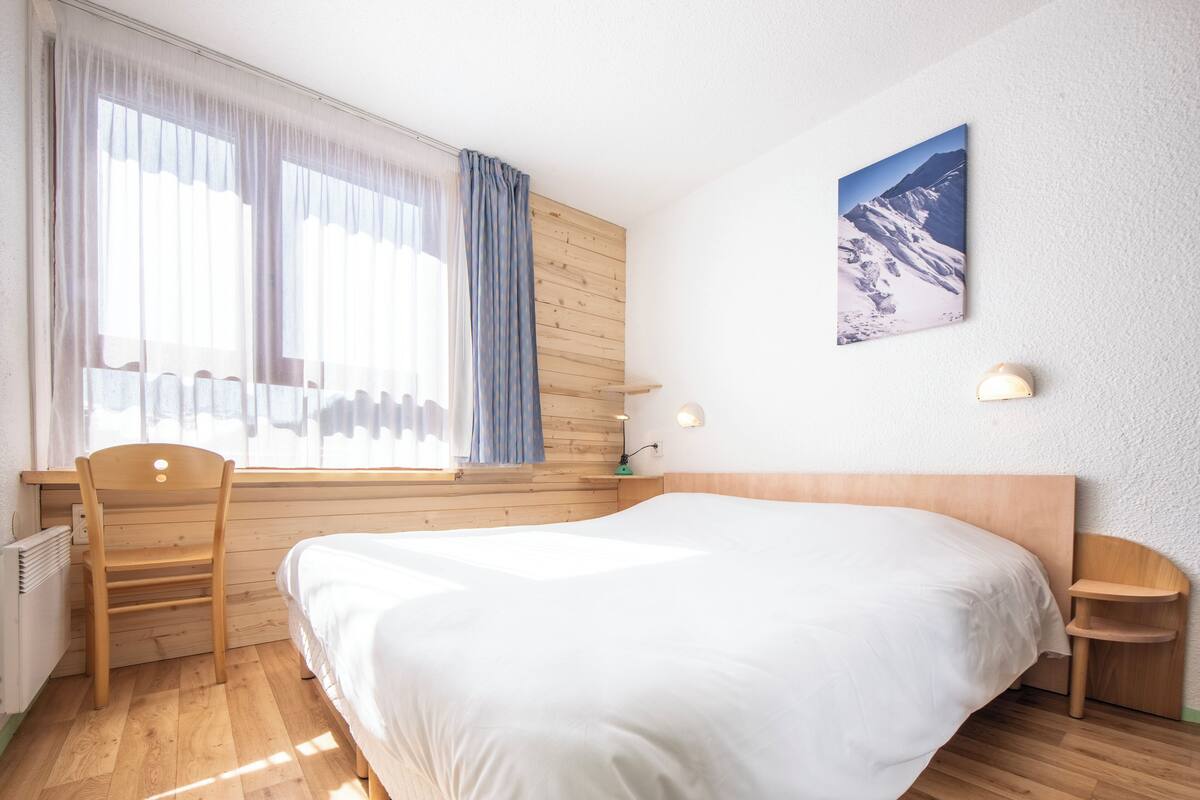 Une chambre à coucher avec une tête de lit en bois, un lit avec une literie blanche, un bureau en bois, une chaise, une fenêtre avec des rideaux, un tableau accroché au mur et une table de chevet avec une lampe.
