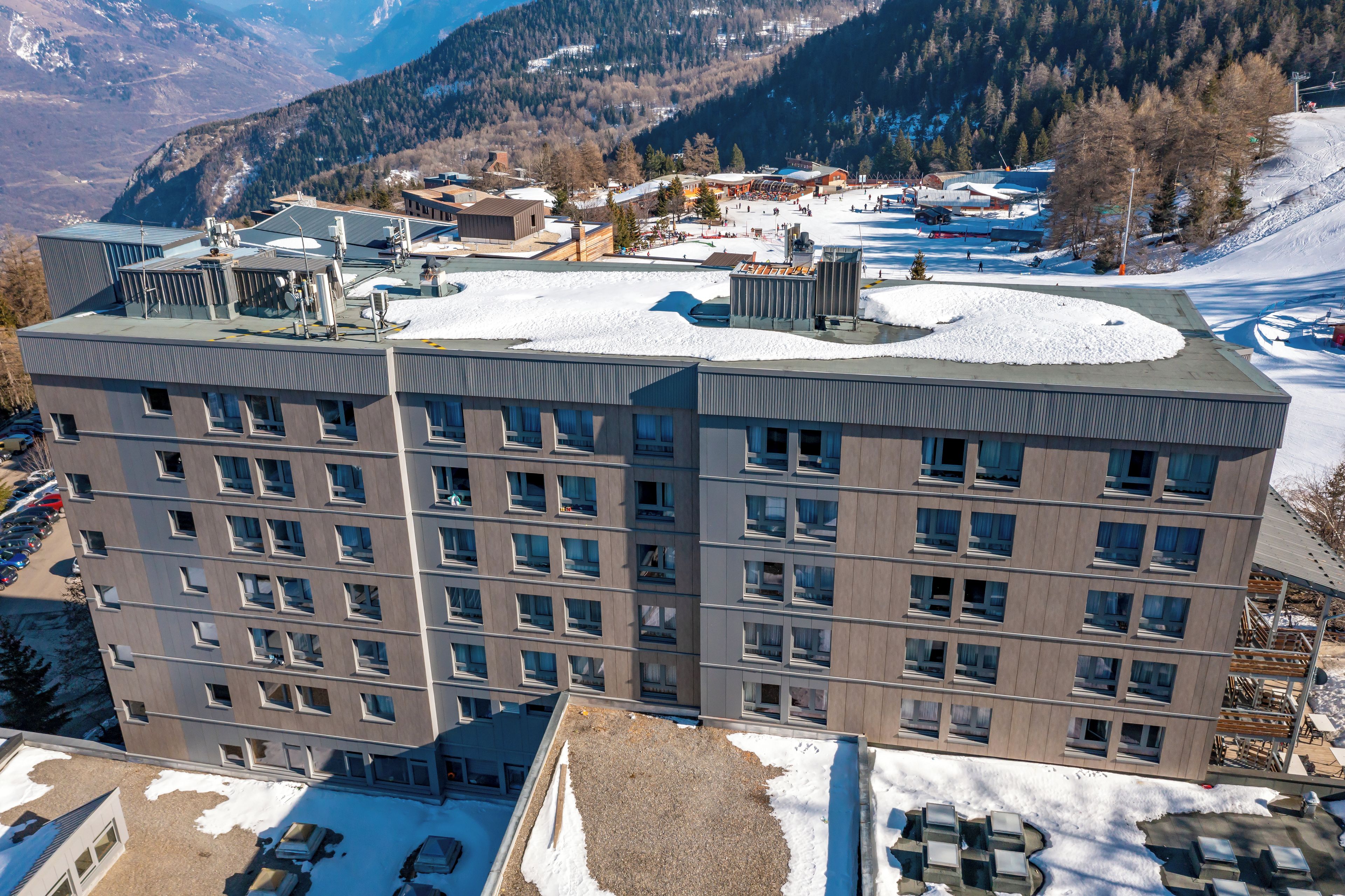 Een meerlaags hotel met een plat dak, grote ramen en uitzicht op een besneeuwd landschap met bergen op de achtergrond.
