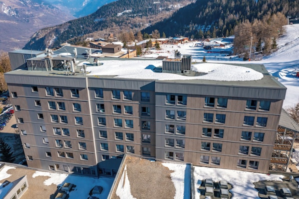 Un hotel de varios pisos con techo plano, ventanas grandes y vistas a un paisaje nevado con montañas al fondo.