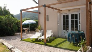 Eine Terrasse mit einer Holz-Pergola, zwei weißen Lounge-Stühlen, einem Tisch mit grüner Tischdecke und einem Blumentopf.