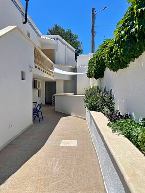 Un patio estrecho iluminado por el sol, con una silla azul, una pared blanca con un diseño decorativo y una maceta con plantas verdes.