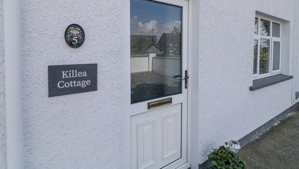 Una casita blanca con una puerta y una ventana. Hay un letrero en la pared que dice "Killea Cottage".