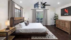 A modern bedroom with a large bed, a ceiling fan, a TV displaying the Netflix logo, and a view of the outdoors through a sliding door.