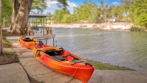 Two kayaks parked by a riverside path with a dock and trees in the background.