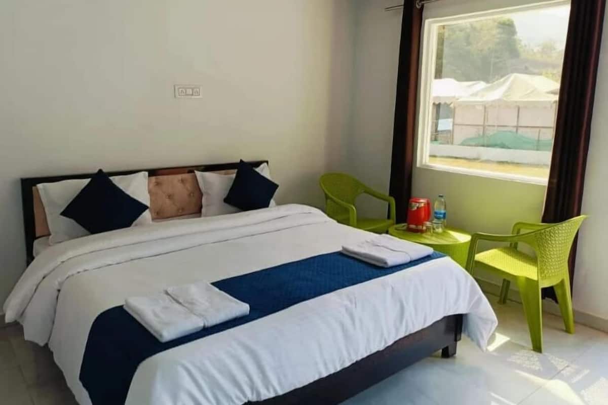 Cama bem arrumada com roupa de cama branca e azul, duas cadeiras verdes e uma pequena mesa com uma garrafa e uma xícara.