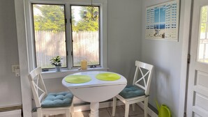 Un petit coin repas avec une table et des chaises blanches, deux fenêtres donnant sur une clôture en bois, un luminaire suspendu et un calendrier accroché au mur.