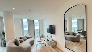 A modern living room with a large curved sofa, a small round table, a flat-screen TV mounted on the wall, and a large mirror reflecting the room. - Manchester Oasis - Modern 3 Bedroom Apartment (Manchester)