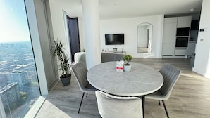A modern dining area with a round table and four chairs, a large window with a city view, a flat-screen TV, and a kitchen in the background. - Manchester Oasis - Modern 3 Bedroom Apartment (Manchester)