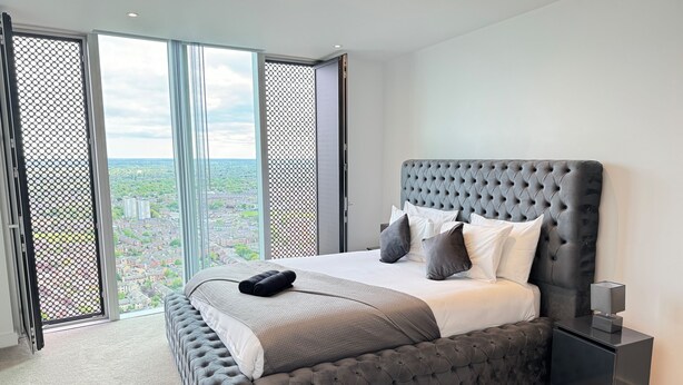 A bedroom with a large bed, grey upholstered headboard, and a view of a cityscape through a sliding door. - Manchester Oasis - Modern 3 Bedroom Apartment (Manchester)