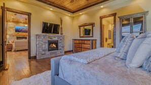 A bedroom with a stone fireplace, a large bed, a ceiling fan, and a TV mounted above the fireplace. - Experience Ruidoso from the top, 360 views,private pickleball, hot tub (Ruidoso)