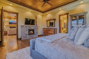 A bedroom with a stone fireplace, a large bed, a ceiling fan, and a TV mounted above the fireplace. - Experience Ruidoso from the top, 360 views,private pickleball, hot tub (Ruidoso)