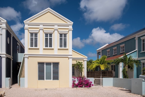 A two-story building with a beige facade, white trim, and shutters. It has a small front porch with a white railing and a pink flowering plant in the foreground.
