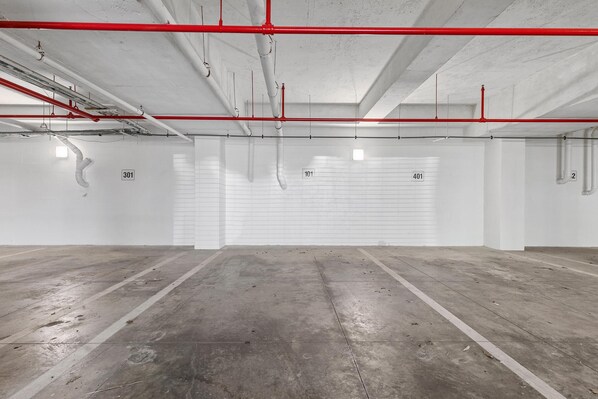 Un estacionamiento amplio y vacío con pisos de concreto y paredes blancas, con tuberías rojas y blancas y puertas de garaje numeradas.