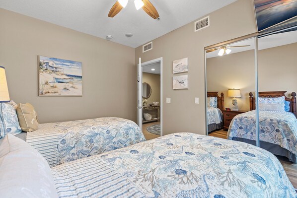 A bedroom with two beds, a ceiling fan, a beach painting, and a mirror reflecting the room. - Atlantic Villas 101 (New Smyrna Beach)