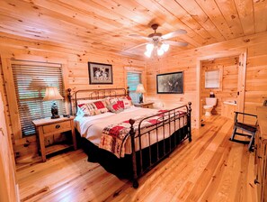 A wooden cabin bedroom with a bed, bedside tables, a ceiling fan, and a bathroom with a toilet and sink.