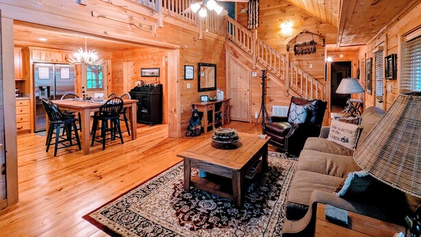 A spacious cabin interior with a dining area, a living room with a sofa and a coffee table, a staircase, and a kitchen with a refrigerator.