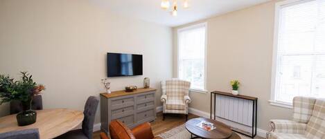 Una sala de estar con un sofá de cuero marrón, una mesa de centro redonda, un sillón a cuadros, un sillón gris, una mesa de comedor de madera con sillas, un televisor de pantalla plana montado en la pared y una lámpara en el techo.