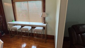 Ein Eckzimmer mit Holzboden, einem Fenster mit Jalousien, einem Schreibtisch mit zwei Stühlen und einer Lampe.
