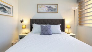 Ein ordentlich bezogenes Bett mit einer gebügelten Kopfteil, zwei Nachttischlampen, ein gerahmtes abstraktes Kunstwerk über dem Bett und ein Fenster mit Jalousien auf der rechten Seite.