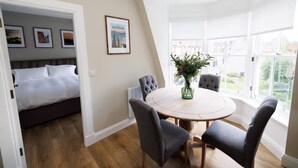 A hotel room with a bed, a round dining table with four chairs, a vase with flowers, and a large window offering a view of the city. - Cosy Apartment in the Heritage Quarter - Sleeps 2 (Dunfermline)