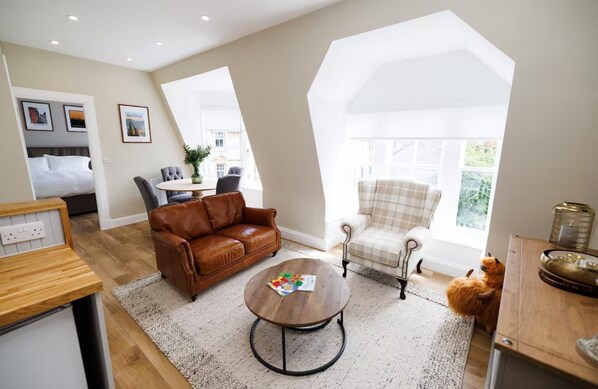 A spacious living room with a brown leather sofa, a plaid armchair, and a round coffee table. There is a dining area with a table and chairs, and a large window providing natural light. - Cosy Apartment in the Heritage Quarter - Sleeps 2 (Dunfermline)