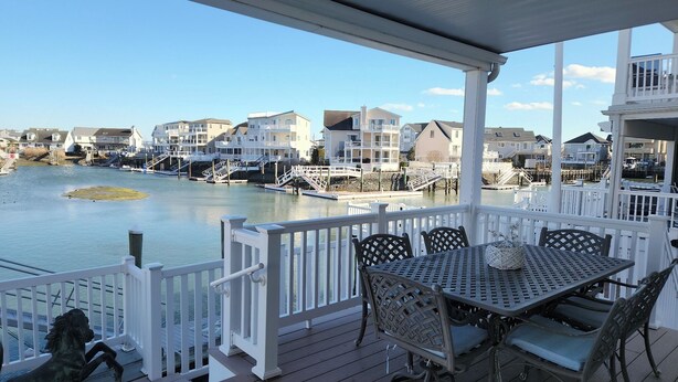Une terrasse couverte, dotée d’une table et de chaises, qui offre une vue sur le bord de l’eau, avec des maisons et une petite île.