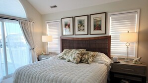 Une chambre d’hôtel avec un grand lit, deux tables de chevet, une petite table, une fenêtre avec des stores et trois tableaux encadrés accrochés au mur.