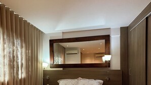 A hotel room with a large bed, a wooden headboard, two bedside tables with lamps, a mirror above the bed, and a window with curtains. - Loft with pool - Near Riocentro and Barra (Rio de Janeiro)