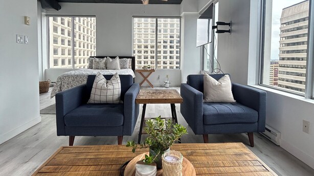 A modern living room with two sofas, a wooden coffee table with decorative items, and a ceiling fan. Large windows offer views of the cityscape. - Downtown Suite - Stunning View Right in the centre of Edmonton (Edmonton)