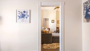 A hallway with a framed floral artwork on the left, a doorway leading to a living area with a sofa and a patterned rug, and a framed picture on the right.