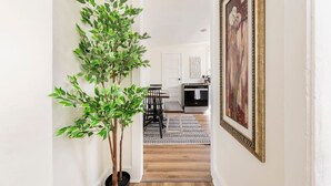 A hallway with a potted plant, a dining area with chairs, a kitchen with cabinets, and a framed picture on the wall.
