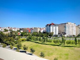 Paisagem urbana com um edifício alto em destaque, área verde com árvores e céu azul claro.