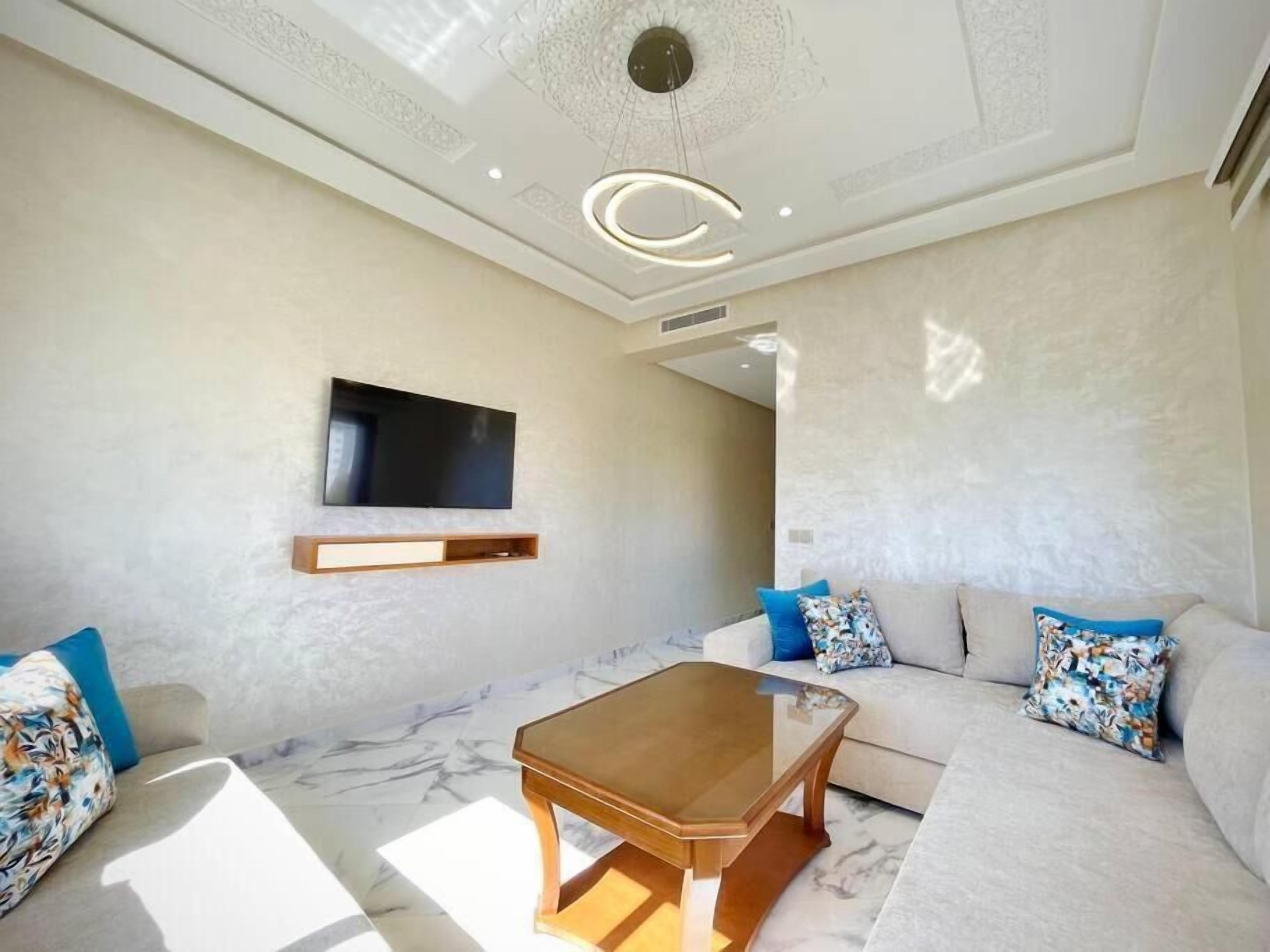 Una sala moderna con un televisor de pantalla plana montado en la pared, una mesa de centro de madera y un sofá color beige con cojines estampados.