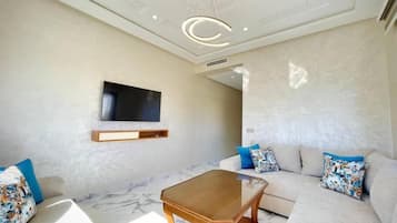 A modern living room with a flat-screen TV mounted on the wall, a wooden coffee table, and a beige sofa with patterned cushions.