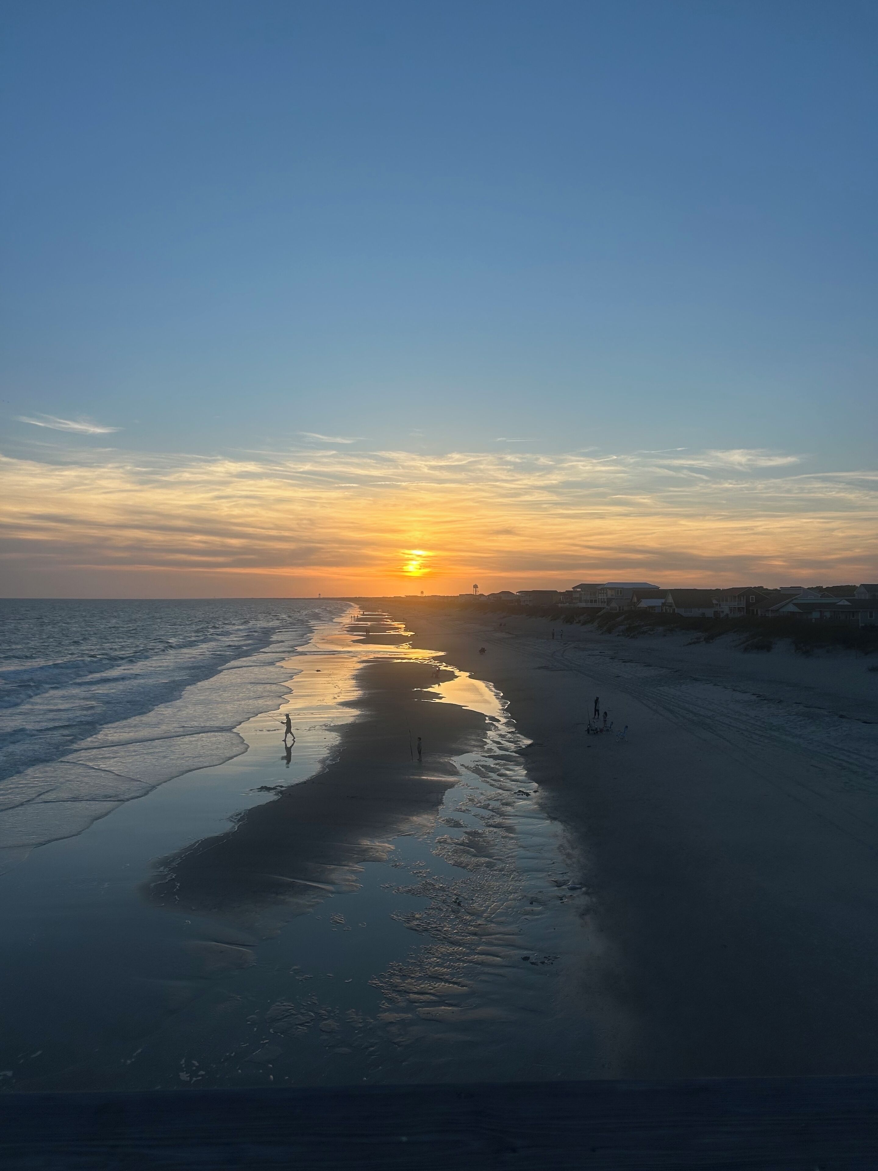 Ein Strand bei Sonnenuntergang mit ruhiger See, wolkenlosem Himmel und Gebäuden in der Ferne.