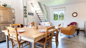 A dining area with a wooden table set for four, surrounded by wooden chairs. A staircase leads to an upper level, and a window provides a view of greenery outside. - Vacation home in Normandy (Aure sur Mer)