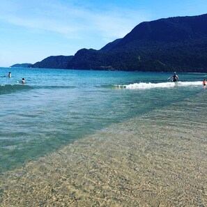 A beach with clear shallow waters, a few people swimming and surfing, and mountains in the background. - Casa c Wi-fi Jacuzzi e Churrasqueira em Guaec SP (São Sebastião)