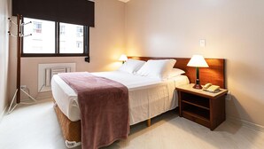 A hotel room with a bed, two bedside lamps, a nightstand with a telephone, and a window with a view of buildings.