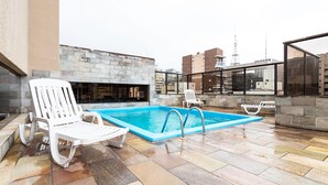 A rooftop pool area with two white lounge chairs, a stone wall, and a view of the city skyline.