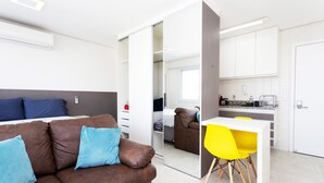 A modern, compact living space with a brown leather sofa, a small dining table with yellow chairs, a kitchenette, and a mirrored closet. - Apartamento com Wi-fi pr Ximo ao Metr Luz (São Paulo)