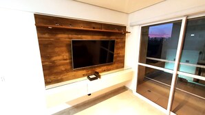 A modern living room with a large flat-screen TV mounted on a wooden wall, a sliding glass door leading to a balcony, and a small wooden shelf above the TV. - Apartamento com Wi-fi pr Ximo ao Metr Luz (São Paulo)