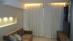 A modern living room with a sofa, a wooden bench, and a large window with curtains. - Apto Beira-mar com Wi-fi e Piscina em Fortaleza (Fortaleza)