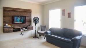 A living room with a flat-screen TV mounted on a wooden panel, a grey armchair, a black leather sofa, and a standing fan. - Casa com Piscina e Wi-fi em Itapetininga (Itapetininga)