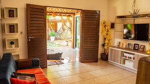 A living room with a sofa, a television, and a door leading to an outdoor area with a hammock and plants. - Casa em Cond Fechado em Barra de Santo Ant nio AL (Barra de Santo Antonio)