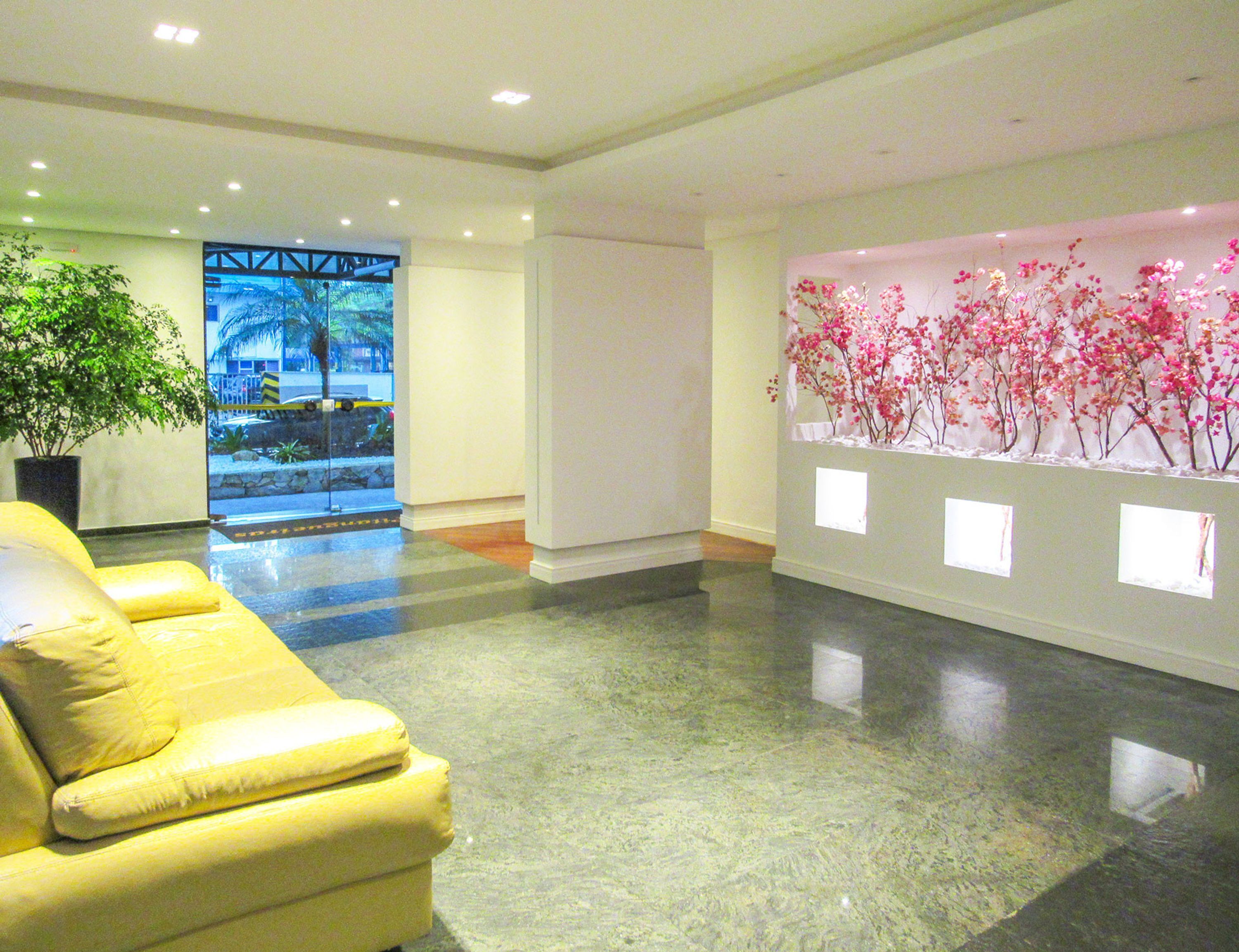 A lobby with a yellow sofa, a large potted plant, a wall with pink flowers, and a view of a street with palm trees.