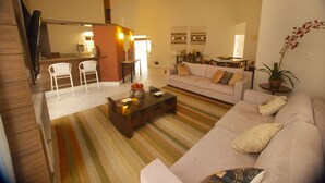 Ein Wohnzimmer mit einer Couch, einem Couchtisch und Sesseln. An der Wand ist ein Fernseher angebracht. Im Hintergrund ist eine Küche zu sehen.